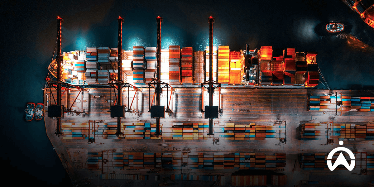 Ảnh chụp từ trên cao một cây cầu với hai xe tải đang băng qua và hai tàu container bên dưới, mô tả hệ thống quản lý đội xe trong logistics đa phương thức.